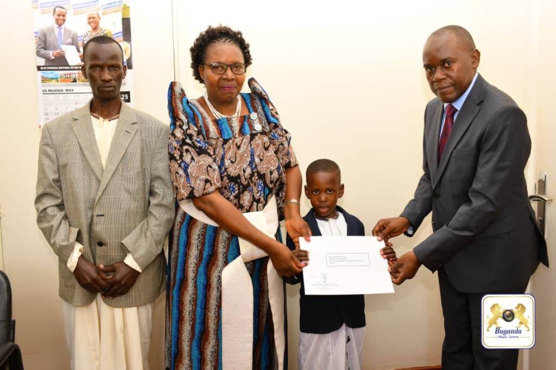 Hon. D.D. Mukiibi, the Kabaka’s Private Secretary, presents the King’s letter of appreciation to Dominic Luther Mubiru, accompanied by his grandmother, at Bulange in Mengo. Hon. D.D. Mukiibi, the Kabaka’s Private Secretary, presents the King’s letter of appreciation to Dominic Luther Mubiru, accompanied by his grandmother, at Bulange in Mengo.